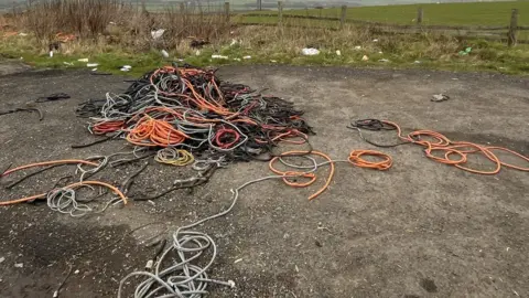 A large amount of cables and cable sleeving coloured orange, grey, black and red have been dumped on a country road. There is other waste that has been dumped in the background. 