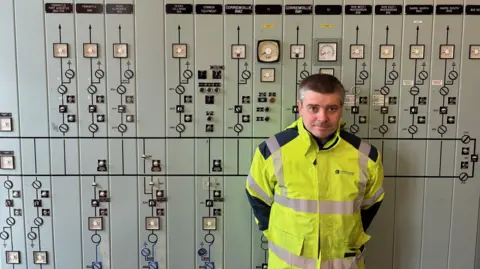 Greg Clarke wearing a high‑visibility yellow safety jacket stands in front of a large industrial control panel filled with dials, gauges, switches, and indicator lights.
