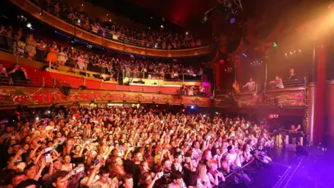 Getty Images A massive, dense crowd of people with upraised hands packing the floor and balconies of a historic, red-lit music hall venue.