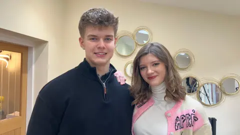 A young man in a black quarter zip jumper is smiling at the camera and standing next to a teenage girl with wavy brown hair who is wearing a white turtle neck jumper and pick and white cardigan with the letters APRA visible on her shoulder