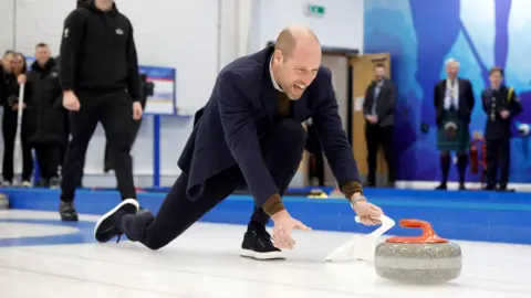 The Prince and Princess of Wales Prince William crouching on a curling rink with his hand on some white plastic equipment. 
