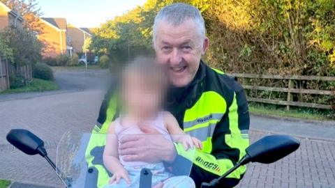 Motorcyclist, Nigel Osler, sat on a stationary motorbike in a driveway, carrying a baby whose face is blurred