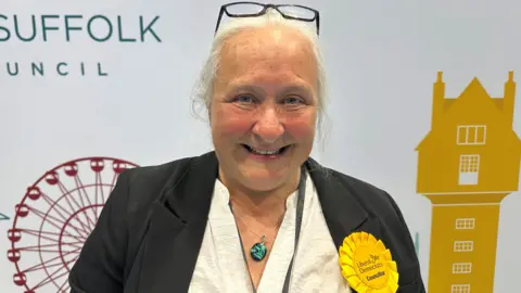 Ben Parker/BBC A head and shoulders image of a woman wearing a black jacket with a yellow rosette pinned to a lapel. She is wearing a necklace with a green pendant and smiling while looking into the camera.