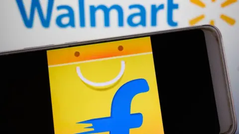 The Flipkart logo - a blue-coloured F against a yellow shopping bag - is seen on a phone screen held against Walmart's blue-and-yellow log