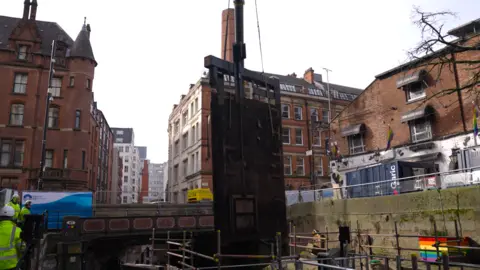 Photograph of the old lock gates being lifted from the Rochdale Canal in Manchester city centre. The image shows a crane, scaffolding and workmen. 