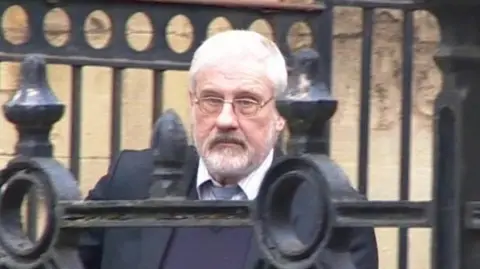 The BBC's Stephen Cox, wearing a button-down shirt and tie, emerges from Reading Crown Court, the railings in front of him as he descends a ramp outside the building. He wears glasses and has short white hair and a white beard.