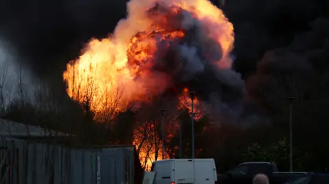 Reuters A huge fireball erupts in the sky behind some trees and metal railings. Traffic is parked in the foreground looking on.