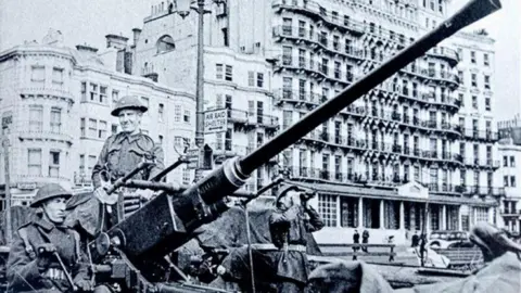 The Regency Society A black and white image of three soldiers and a large anti-aircraft gun in front of a large hotel. 