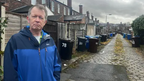 Stuart Atkinson stood in an alley way with lines of wheelie bins the background on a cobbled path
