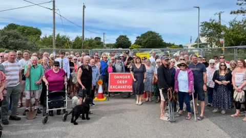A picture of a group of residents looking forward. They are stood in front of a metal fence with a sign that reads 'Danger Deep Excavation' on it.