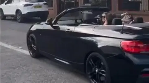 Hannah Taylor Screen grab of Hannah Taylor, wearing sunglasses, smiling as she drives a teenage girl and boy who are in the rear seats of her open-top convertible