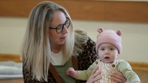 A woman with long blonde hair holds a baby in her arms