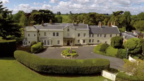 Moulton Grange is a cream coloured building with a grey slate roof and brick chimneys. This view shows the front façade, including an entrance porch with two columns on either side, a bay window to the left of the porch and multiple lattice pane windows. The approach to the property is a wide driveway surrounded by hedges with a circular bush in the centre of the drive and a union flag on a white pole.