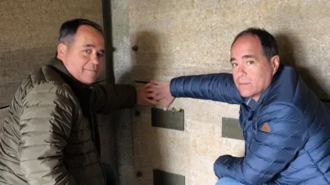 Xavier Lluch Two identical spanish twins in their 50s hold a memorial plaque dedicated to their grandfather