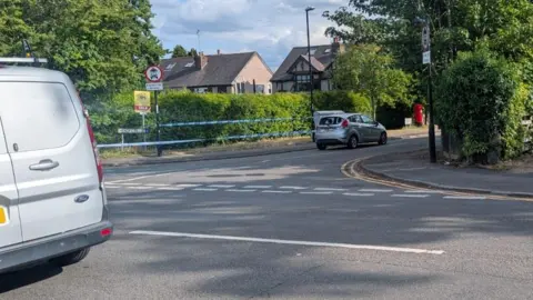 Police tape runs across a road junction. The back of a grey van can be seen near to the camera. Further away, with some of the police tape around it, is a silver car. Behind that is a hedge and homes.