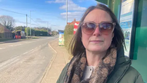 Matt Precey/BBC Woman in dark glasses standing at a bus stop. She is wearing a green coat. There is a road beside her. 