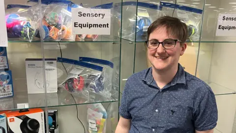 BBC A person in a blue short-sleeved shirt stands smiling in front of a glass cabinet filled with noise cancelling headphones and fidget toys. A sign reading 'Sensory Equipment' is taped to the front of the cabinet.