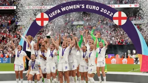 PA Media England women's team in white strip holding their arms aloft and cheering. There is a purple arch above them saying "Uefa women's Euro 2022). They are on a football pitch and there is a crowded stand behind them. 