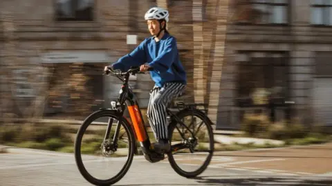 SYMCA A woman wearing blue and white striped trousers, a blue sweatshirt and a white cycle helmet is pictured riding a black and orange electric bike.