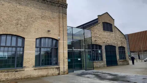 The outside of Sand Martin House in Peterborough. There are two yellow bricked buildings, with a glass atrium in the middle
