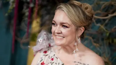Colleen Hoover at a film premiere with her blonde hair in an up-do, wearing dangly white feather earrings - she has a one-shoulder jewelled white dress on, and a tattoo of writing can be seen on her shoulder 