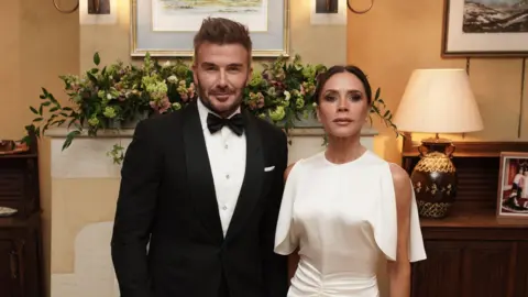 Getty Images David and Victoria Beckham pose side-by-side in front of a mantelpiece, wearing a tuxedo and white dress respectively.