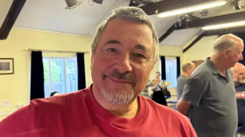Martin Heath/BBC A person wearing a red T-shirt stands in the foreground of a community hall or meeting room. Several other people are visible in the background, standing and talking near rows of chairs. The room has cream-coloured walls, exposed dark ceiling beams, large windows with dark curtains, and bright overhead lighting.