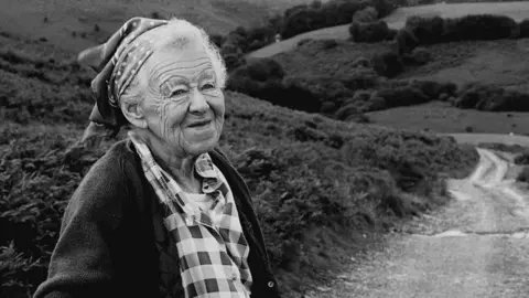 Mark Rattenbury A black and white photograph of an elderly lady with white hair wearing a head scarf, a checked blouse and cardigan, with a road through Exmoor behind her