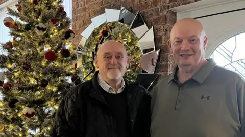 Mia Brannigan/BBC Graham Cutler and Barry Hunt are smiling stood in front of a Christmas tree decorated with baubles. They are both bald men.