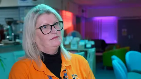 Cheryl Simpson, chief executive of Space4Autism, has bobbed grey hair and wears black glasses. She is wearing a bright orange, open-necked shirt on top of a black pol-necked top.