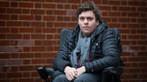 Emma Lynch/BBC Alex Taylor, a white man wearing a black jacket and grey scarf and in a wheelchair, on a London street