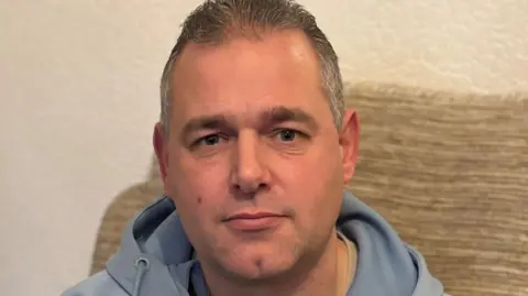 BBC A man in a light blue tracksuit top with short hair sitting on a beige sofa looks at the camera unsmilingly 