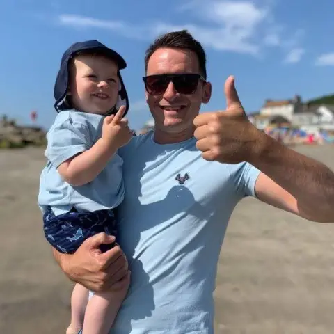 Family handout Adam Keane is wearing a sky blue top and sunglasses, he is holding his thumb up. He is holding his son who is also wearing a light blue shirt, dark blue shorts and hat. 