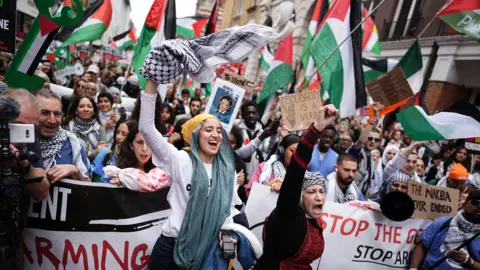 PA Dozens of demonstrators are pictured amongst a sea of Palestinian flags waving in the air. Some are pushing their fists up in the air, and many are holding placards  