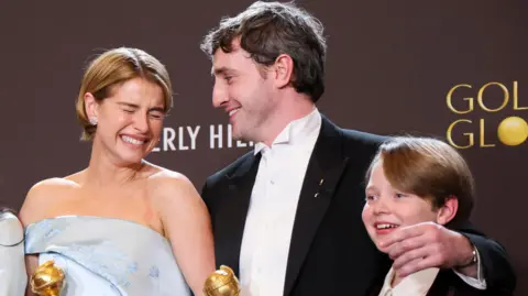 Reuters Jessie Buckley, Paul Mescal e l'attore bambino Jacobi Jupe posano con il premio per il miglior film ai Golden Globes. Buckley, che ha i capelli corti e biondi e un abito argento/azzurro con spalle scoperte, sorride ampiamente con gli occhi chiusi. Mescal ha i capelli corti e scuri e indossa un abito nero e una camicia bianca. Jacobi Jupe ha i capelli corti e biondi e sorride.