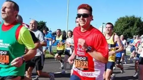 Kristian Shearan Kristian is pictured among a group of runners, wearing a red running t-shirt, as he competes in the Great North Run. He is wearing dark glasses and is smiling as he runs. There are a number of runners around him and it is a bright, sunny day.