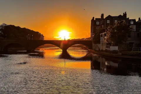 Paul Steven Sunset over a bridge over a river, the sun is reflected on the water.