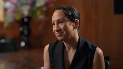 Gleit wears a sleeveless black blazer, and is sitting on a chair in front of a large glossy wood panelled room. A large vase of colourful flowers is also blurred in the background behind her.