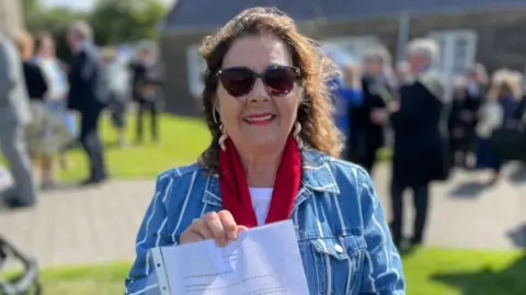 Millie Blenkinsop-French wears a blue and white denim jacket, red scarf and sunglasses. She has curly brown hair. She holds up a white document in a plastic wallet. 