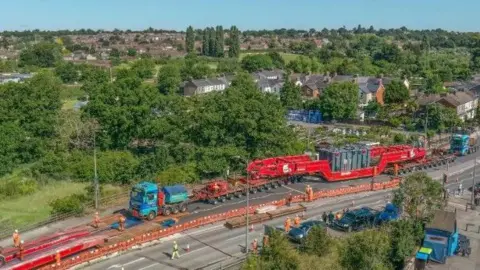 Jeff Welch A large load being carried on the back of a blue truck travels down a road, while workers dressed in orange watch on. 