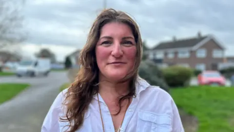 A woman with long brown hair and a white blouse in front of a blurred street with grass verges