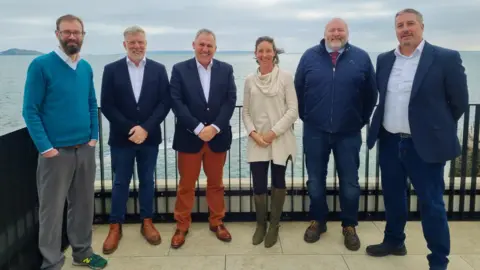 A group of men and women smiling infront of a blue sea and grey sky.