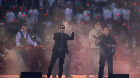 Getty Images Five members of UB40 on a stage in the centre of a stadium, with dry smoke blurring the image.