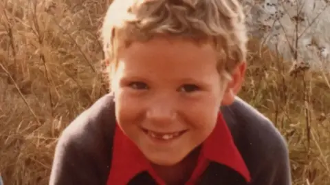 PA Media A younger Hamish Cooper smiles for a photo in a field. He is missing some baby teeth and has blond, short wavy hair, wearing a blue jumper over a red polo shirt.