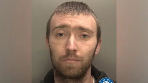 A mugshot of a man with short, brown hair and a beard. He is staring into the camera and wearing a black hoodie.
