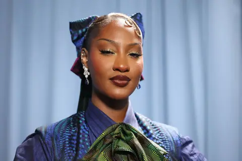 MIKE COPPOLA/GETTY IMAGES A close up of Tems who is wearing a blue and green African-style outfit.