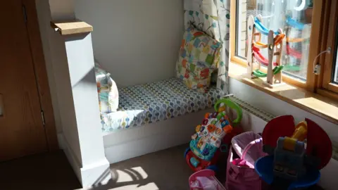 Shaun Whitmore/BBC A small padded sofabed by a window. There are pillows and lots of toys for small children.