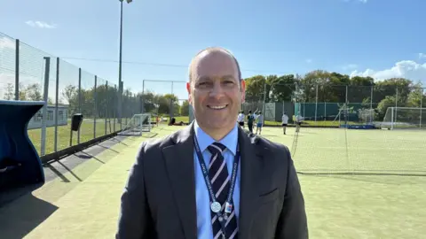Cockermouth School head teacher Richard King standing on the cricket pitch at the school. He is wearing a grey suit jacket, blue shirt and striped tie.
