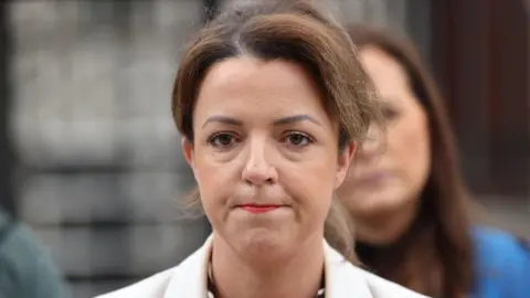 Liz Kimmins has brown hair tied back, has brown/green eyes and is wearing a white suit jacket
