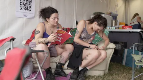 Two women sat on a sofa in the Repairium. The woman on the left is reading a book while the woman on the right is sewing up a ripped skirt.